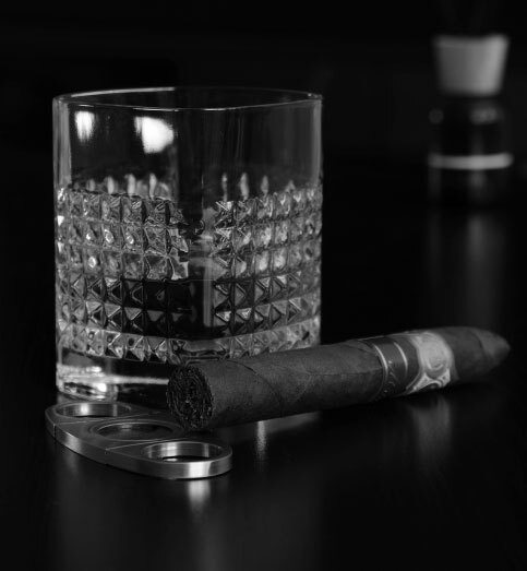 black and white photo of a glass of whiskey on the table with a cigar next to it